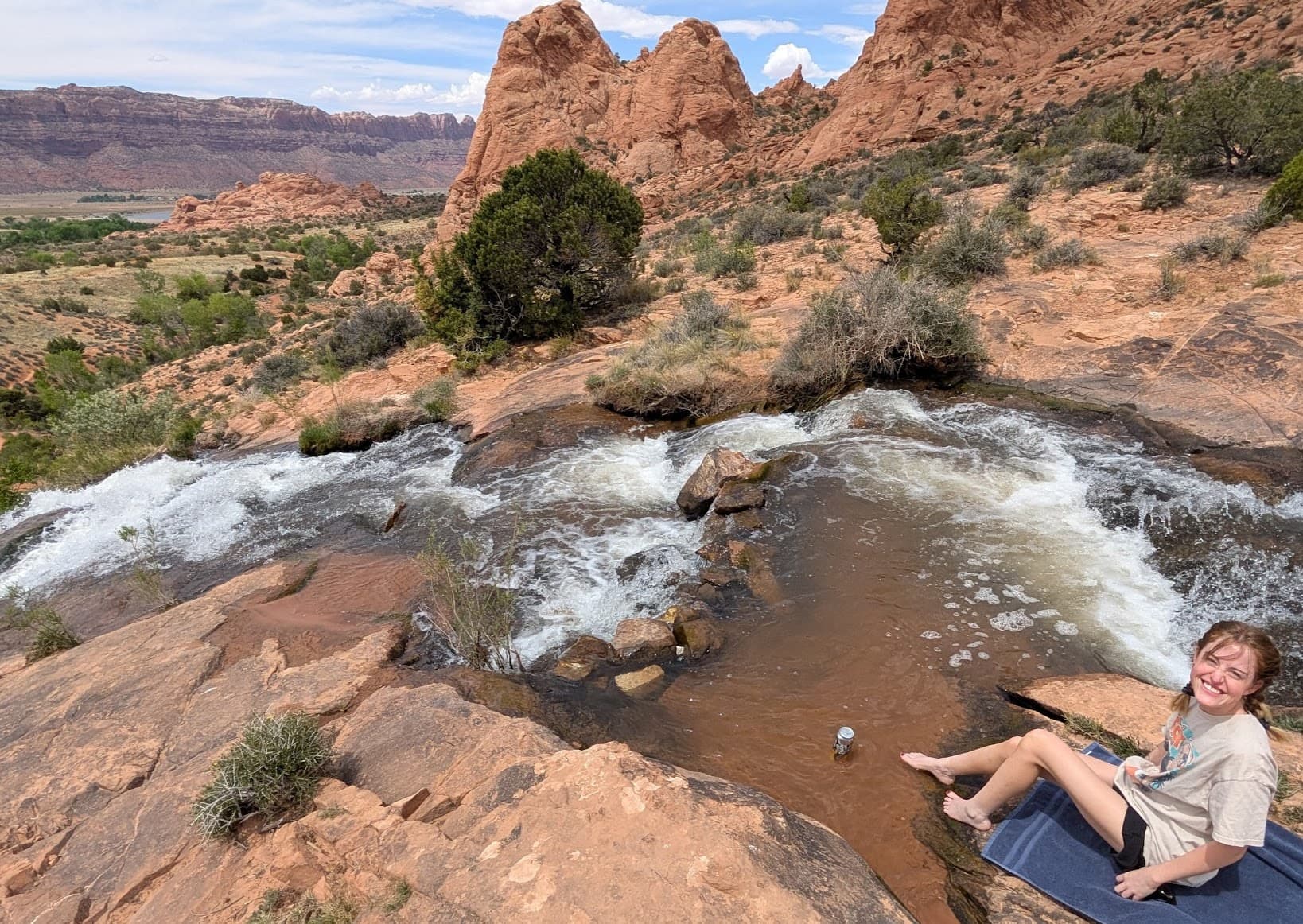 High in a red-rock desert, a small ribbon of water spills over sun-warmed stone, carving a secret pool amid juniper and open sky. An artist rests at the edge, feet cooling in the rush, letting the canyon’s hush and the laughter of water rinse the dust from the day. The moment feels spacious and unhurried, a quiet communion where time slows and the land breathes you back into yourself.