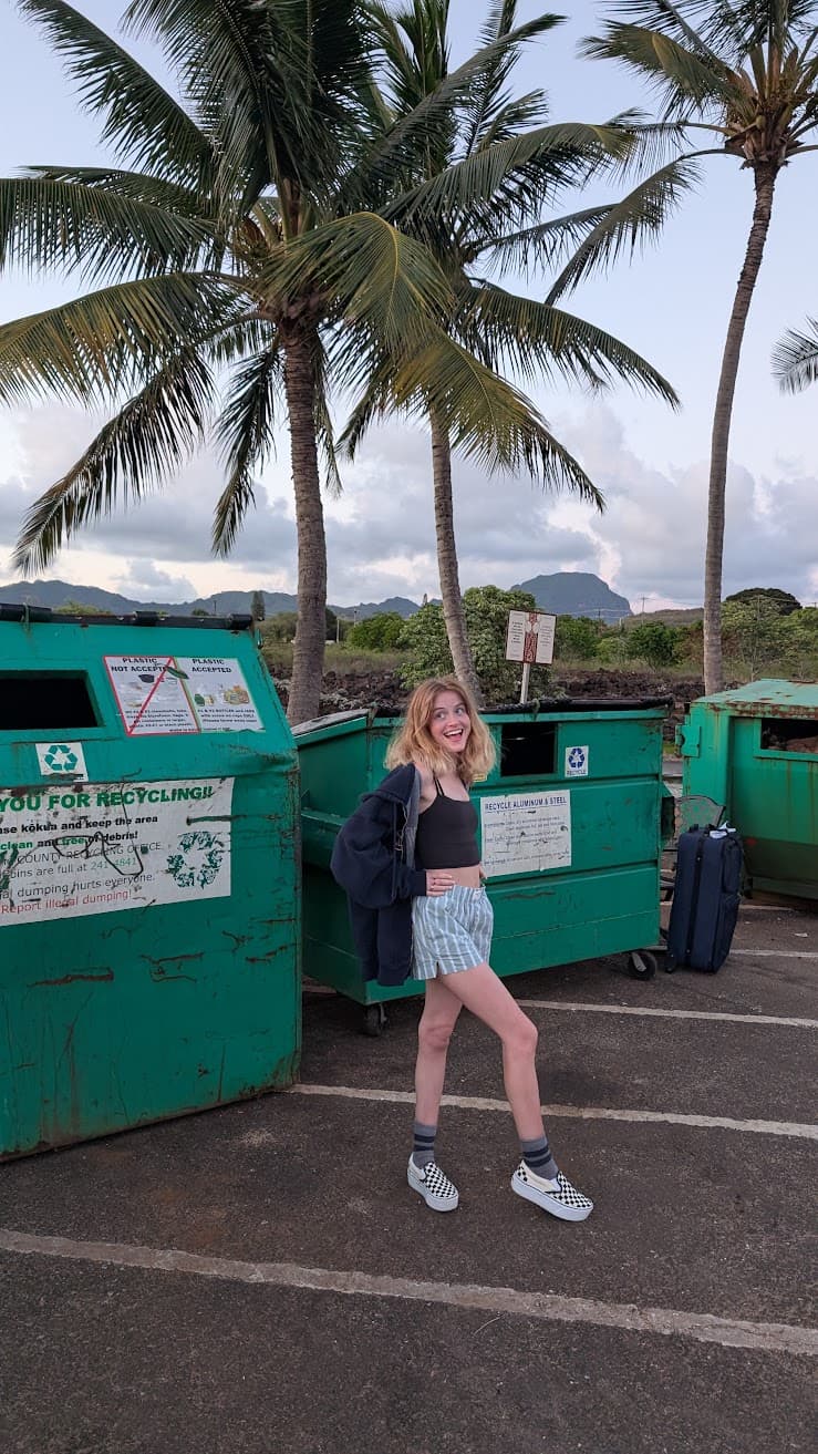 In a sun-warmed parking lot near the sea, palms sway above weathered green bins while distant mountains soften under a pastel sky. A pause between journeys unfolds here—bare legs dusty from the road, a suitcase waiting nearby, laughter rising like a sketch made in the margins of travel. The mood is light and playful, a fleeting moment where art and wandering meet, alive with salt air and the promise of wherever comes next.