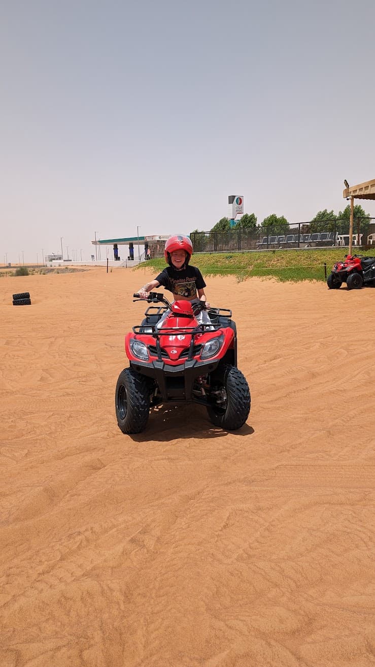 Under a wide, bleached-blue sky, a red quad bike hums across soft desert sand, its tires sketching playful lines where the wind has already been at work. A helmeted rider pauses mid-journey, laughter caught in the heat shimmer, savoring the freedom of motion and the thrill of learning the land by touch and sound. The moment feels sun-warm and weightless, a bright breath of joy between dunes where curiosity and courage meet.