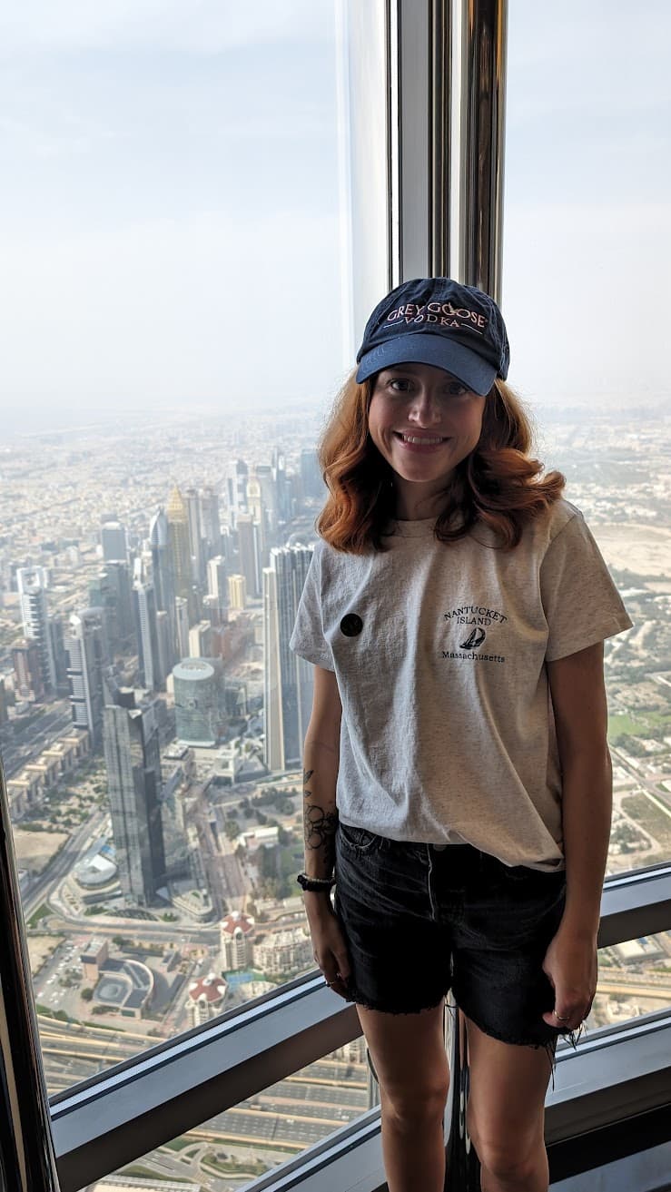 High above a sun-washed city, inside a glass-walled tower, the world falls away into a lattice of streets and shimmering towers, heat-hazed and vast. A quiet pause unfolds here—standing at the window, sketchbook spirit open—where the hum of the city softens into wonder and the moment feels weightless, suspended between sky and earth. The mood is bright and reflective, a gentle thrill of being small inside something immense.