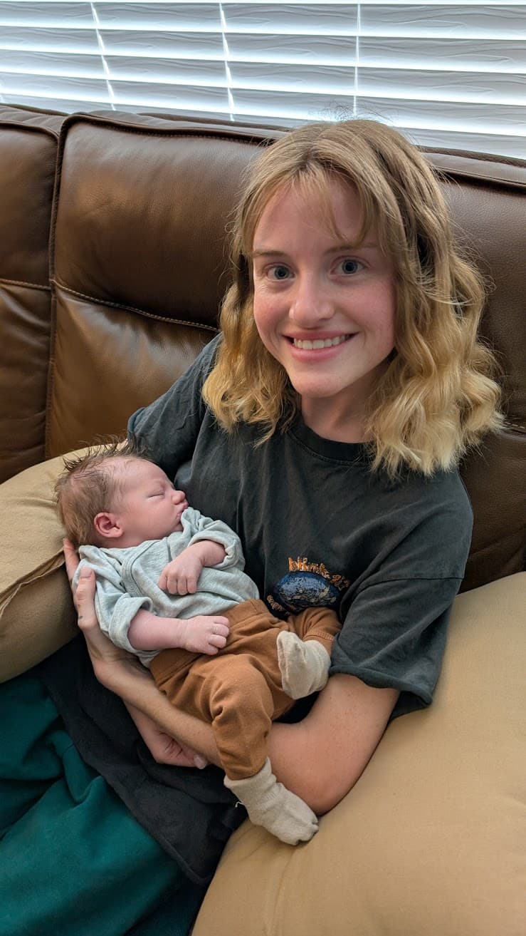 In a quiet living room washed with afternoon light slipping through the blinds, an artist pauses on a well-worn leather couch, cradling a sleeping newborn as if holding a fragile dream. The world feels hushed here—breath slow, time softened—where creation isn’t on canvas or page but nestled warm against the heart. The mood is tender and grounding, a gentle reminder that some of the deepest art is born in stillness.